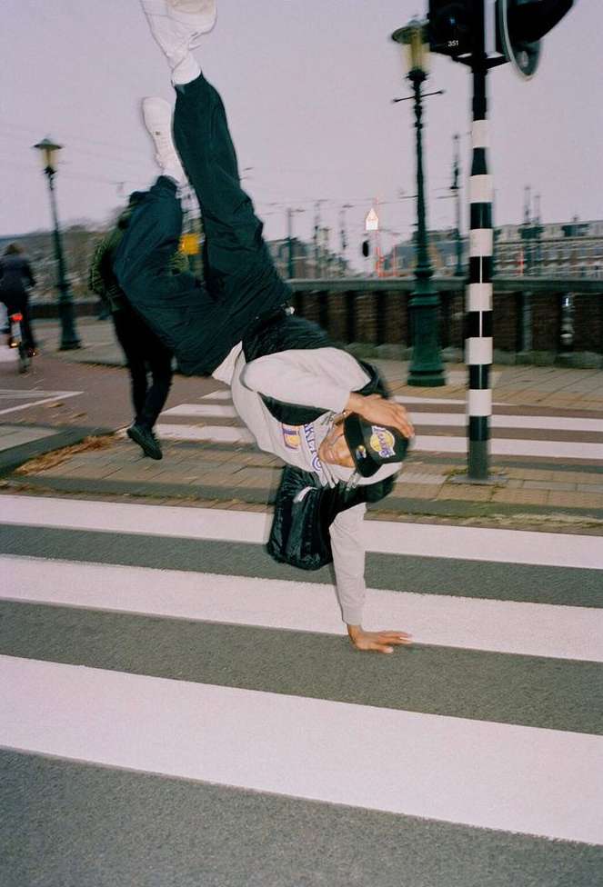 Young adult doing a handstand on a crosswalk in an urban environment, wearing a cap, with other pedestrians in the background.
