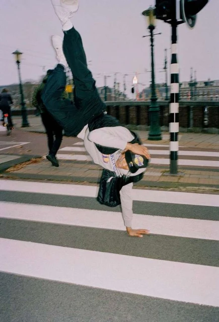 Young adult doing a handstand on a crosswalk in an urban environment, wearing a cap, with other pedestrians in the background.