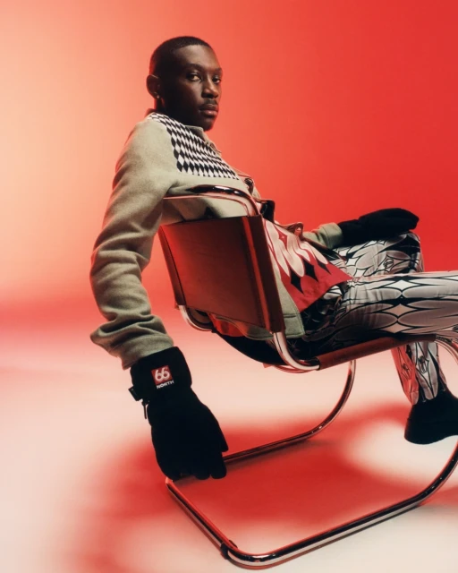 An adult Black male sits on a metal chair in a light grey sweater, patterned pants, and black gloves, posing against a red backdrop.