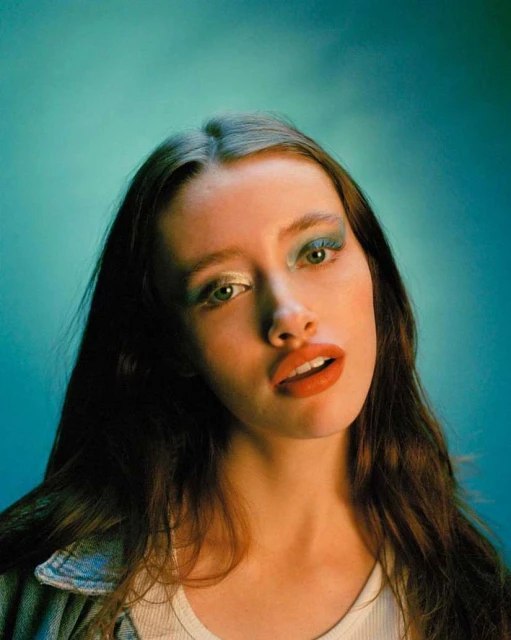 Portrait of a young woman with blue eye shadow and wavy brown hair looking at the camera against a blurred blue background.