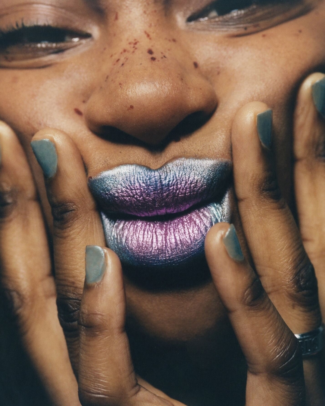Close-up of an adult woman with creative metallic silver and purple lip makeup touching her face with manicured hands and a ring.