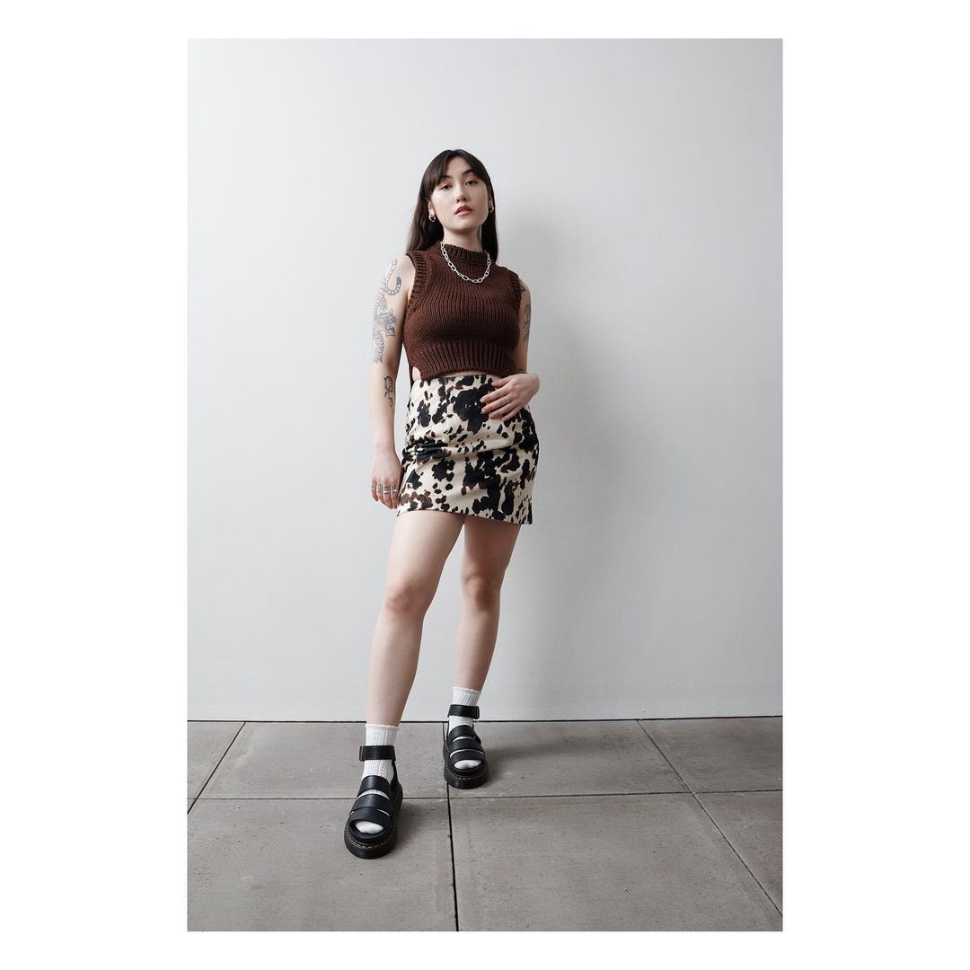 Young woman in a brown knit top and cow-print skirt poses in a neutral studio, complemented by black platform sandals and a delicate necklace.