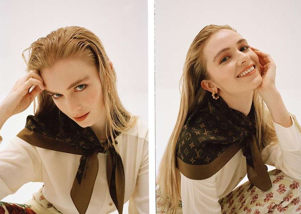 A woman with long blonde hair smiles wearing a brown patterned scarf, a white button-down shirt, and floral pants in a photo studio.