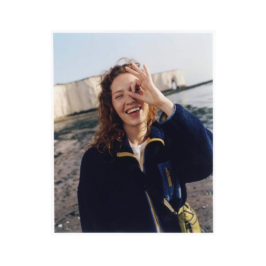 Happy young woman smiles with an OK sign near her eye, wearing a navy fleece jacket at the beach with white cliffs in the background.