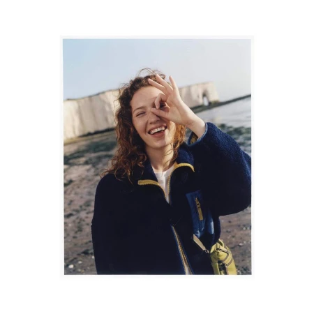 Happy young woman smiles with an OK sign near her eye, wearing a navy fleece jacket at the beach with white cliffs in the background.