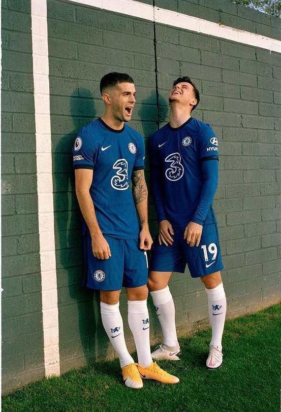 Two soccer players in blue Chelsea uniforms pose against a textured gray wall with grass at the bottom, showcasing team pride.