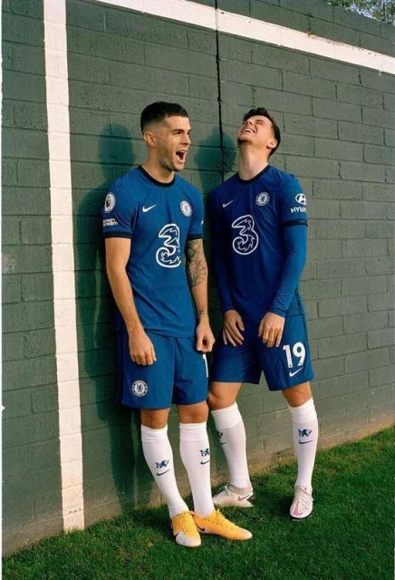 Two soccer players in blue Chelsea uniforms pose against a textured gray wall with grass at the bottom, showcasing team pride.