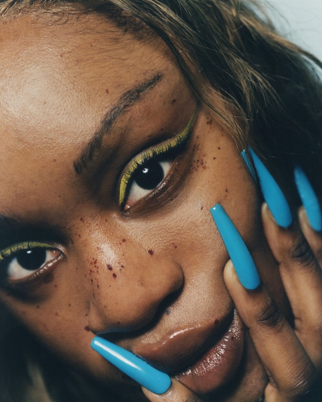 Close-up portrait of a woman with yellow eyeliner, blue manicure, fair skin and brown eyes in a beauty headshot.