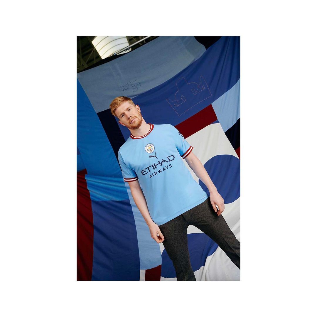 Adult male in a blue Manchester City soccer jersey, standing against a backdrop of blue and white fabric, looking at the camera.