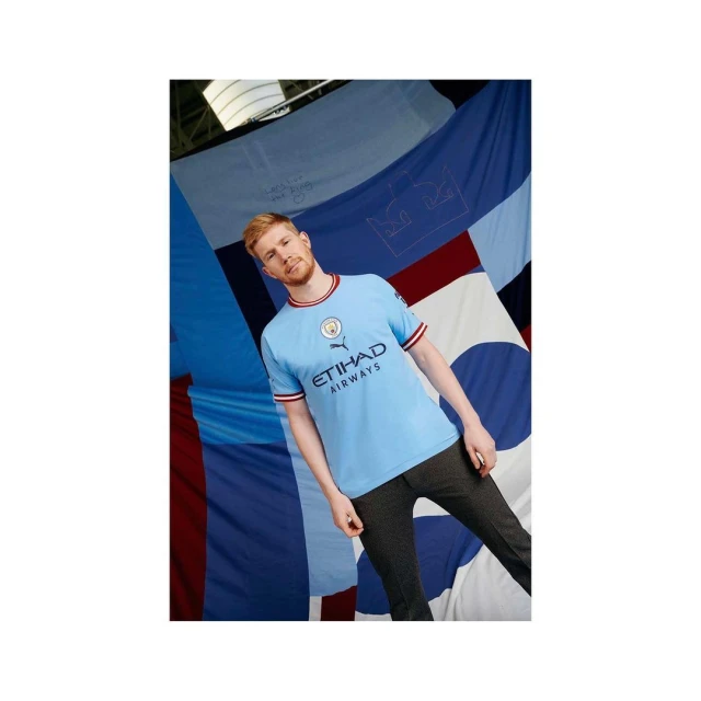 Adult male in a blue Manchester City soccer jersey, standing against a backdrop of blue and white fabric, looking at the camera.