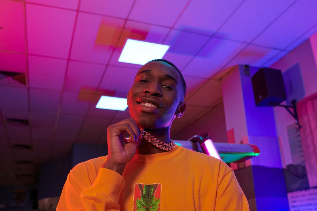 Young man with gold chain poses indoors under vibrant purple and pink lights, wearing a yellow shirt with a graphic design.