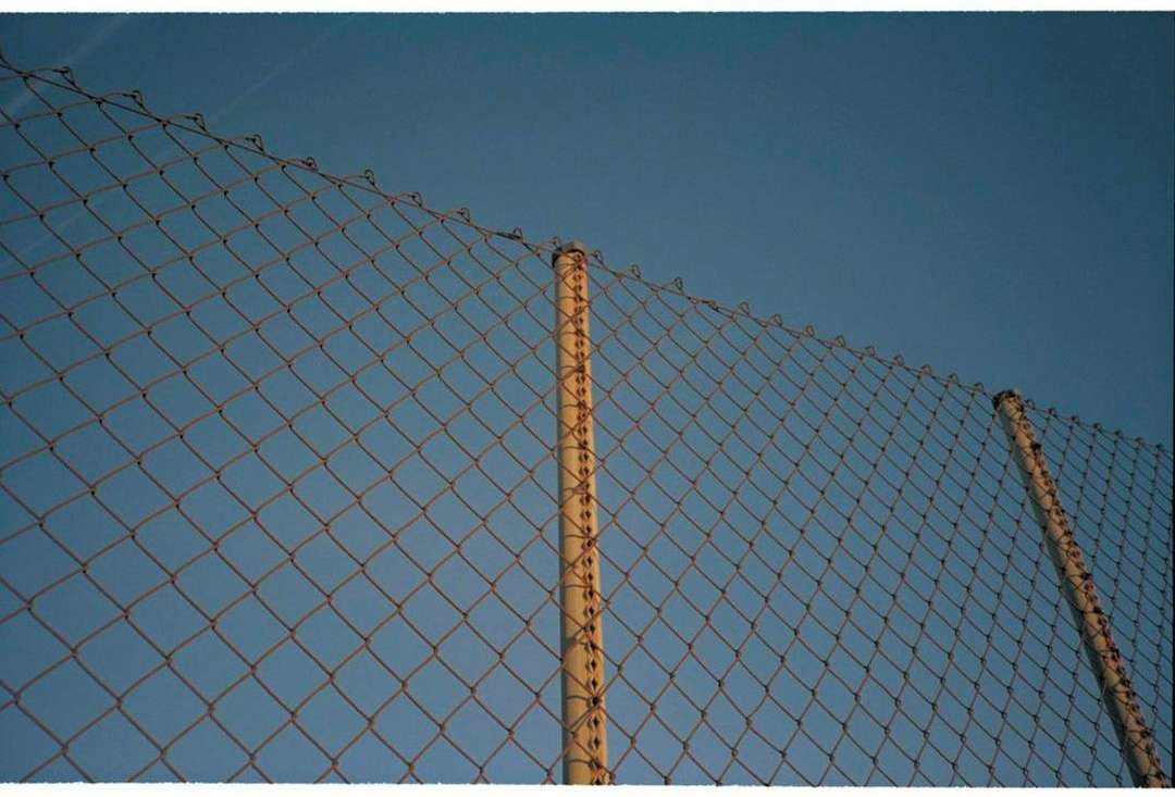 Chain-link fence with steel poles against a clear blue sky, shot from a low angle emphasizing texture and geometric pattern.