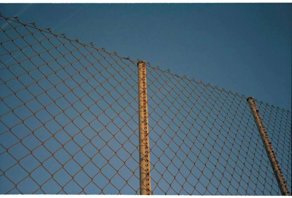 Chain-link fence with steel poles against a clear blue sky, shot from a low angle emphasizing texture and geometric pattern.