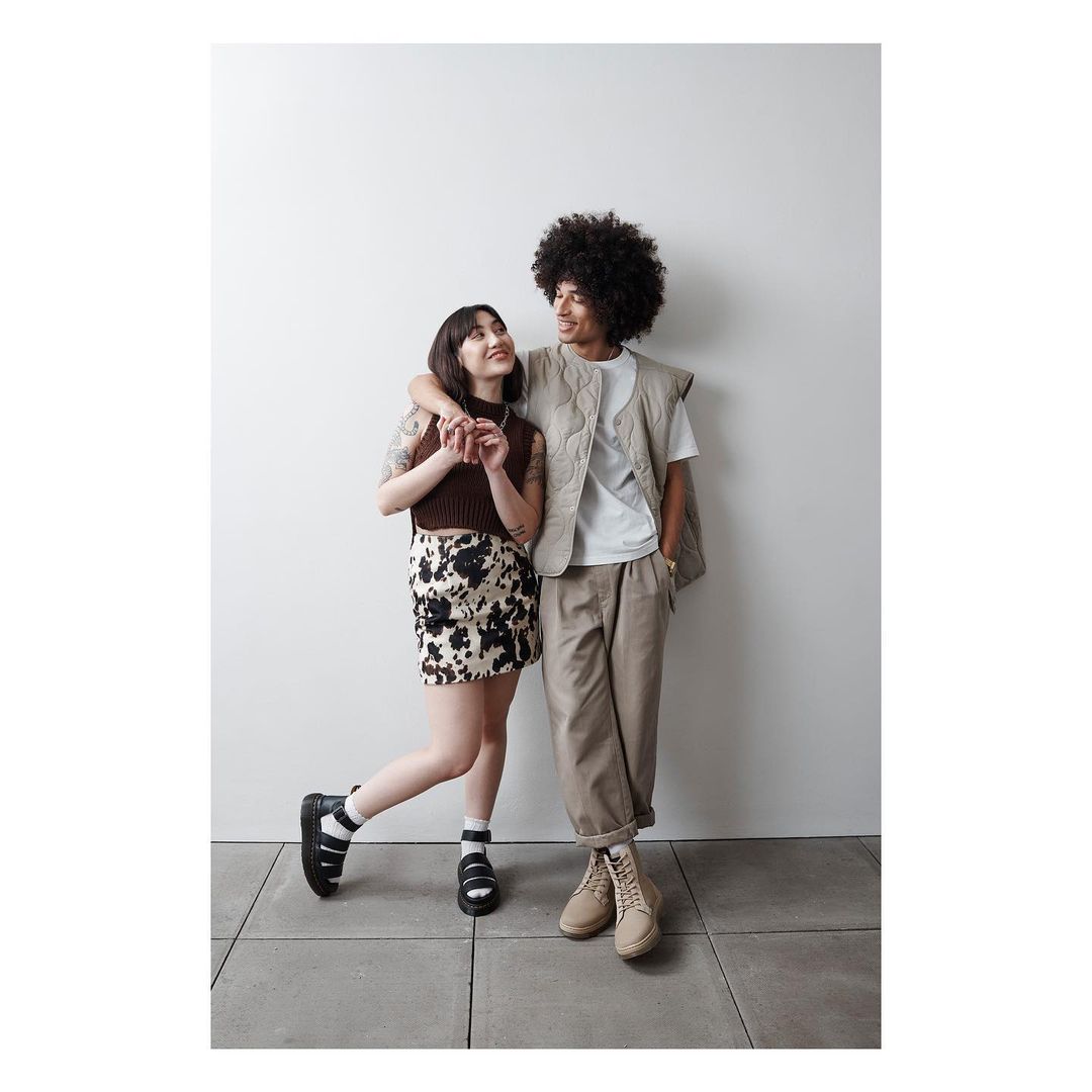 A stylish couple poses indoors with their arms around each other. The woman wears a short skirt and black sandals, while the man has on khakis.