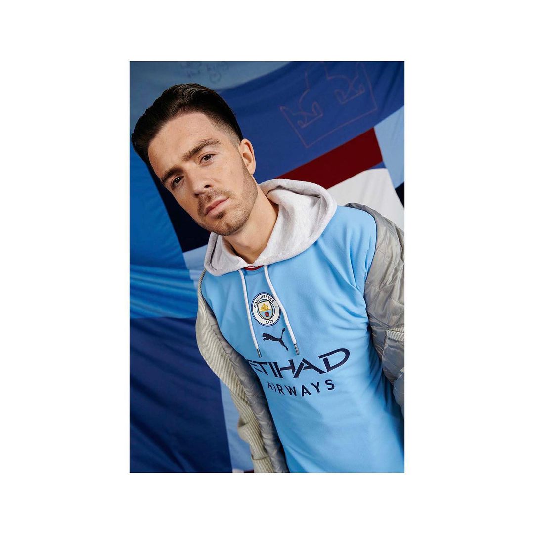 Close-up of a handsome adult male with dark hair wearing a light blue soccer jersey with sponsor logos, posing in a studio setting.
