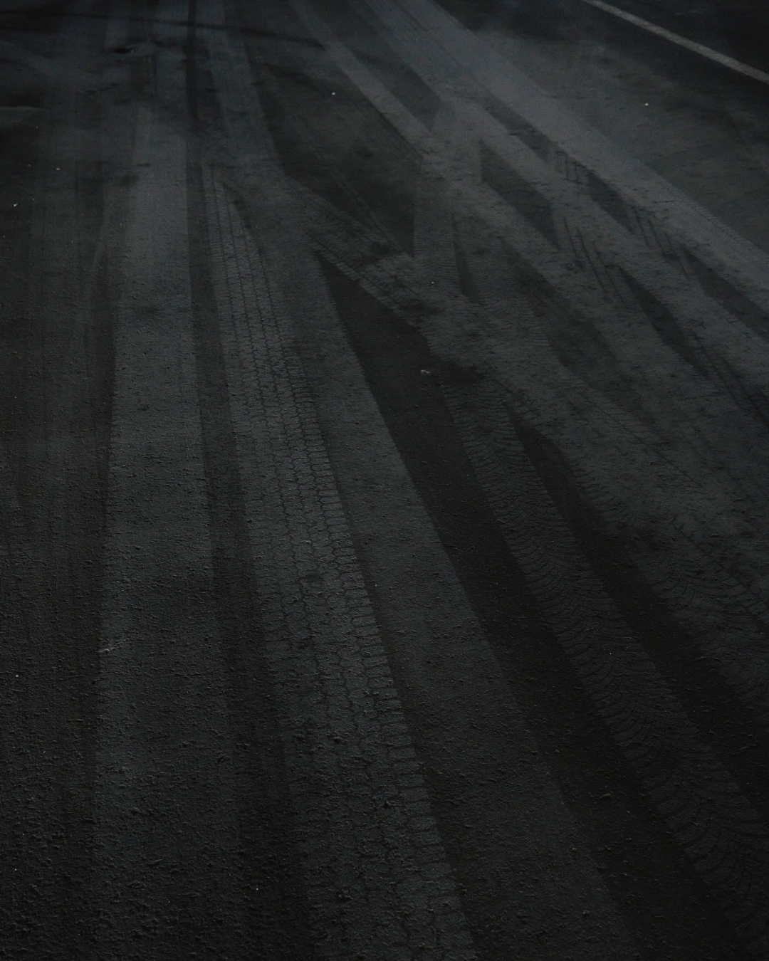 Black asphalt road surface shows clear tire tracks creating textured linear patterns, emphasizing transportation and road usage at night.