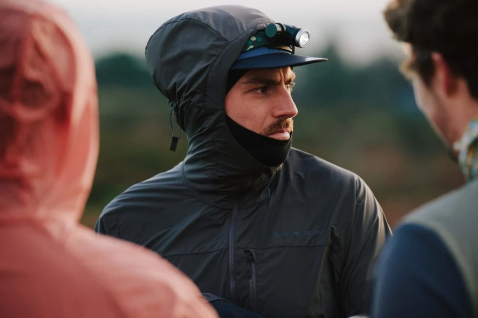 Close-up of adult male wearing a hooded grey jacket and headlamp, standing outdoors with two people in the blurred foreground.
