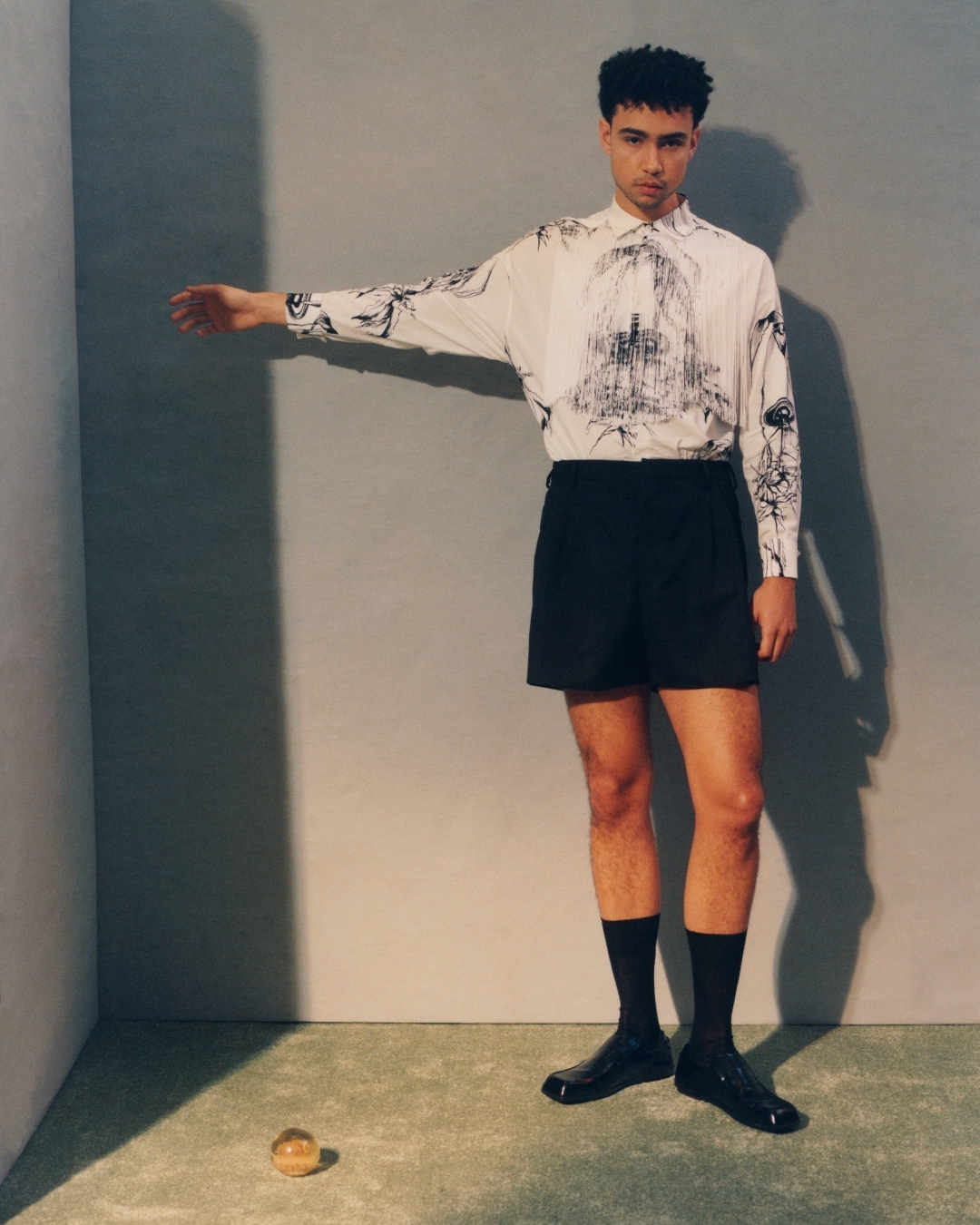 An adult man in a printed shirt and shorts standing indoors with one arm extended, in a studio setting with a clear ball on the floor.