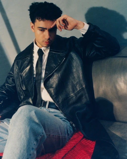 A young adult male poses in a leather jacket, shirt, and tie while seated. The background is a subdued gray and blue color palette.