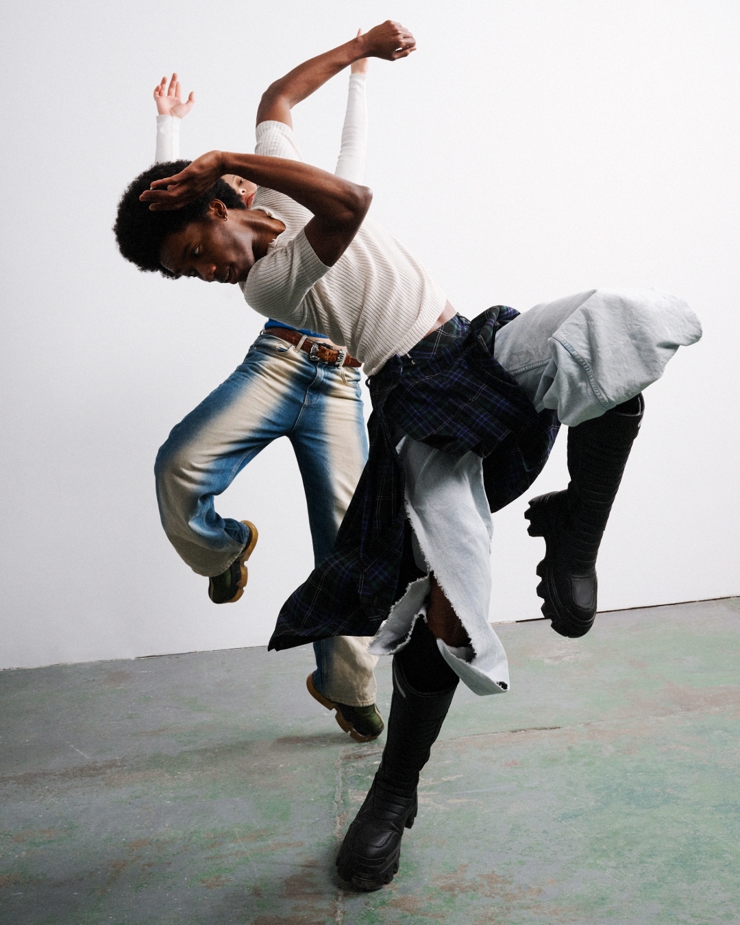 Two African American adults in fashionable clothes perform a dynamic, tilted dance pose against a simple white wall in a modern photo studio.