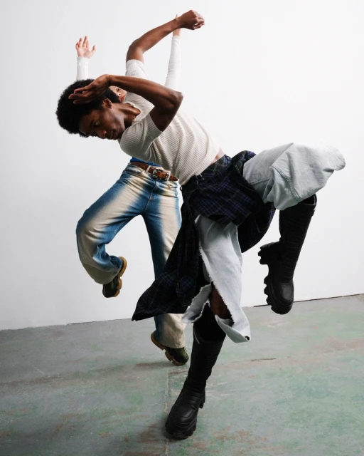 Two African American adults in fashionable clothes perform a dynamic, tilted dance pose against a simple white wall in a modern photo studio.