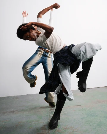 Two African American adults in fashionable clothes perform a dynamic, tilted dance pose against a simple white wall in a modern photo studio.