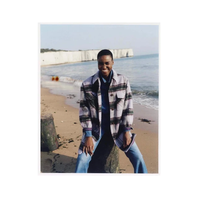 Smiling African woman in a plaid shirt poses on a sandy beach with rocky cliffs and ocean view on a sunny day outdoors.