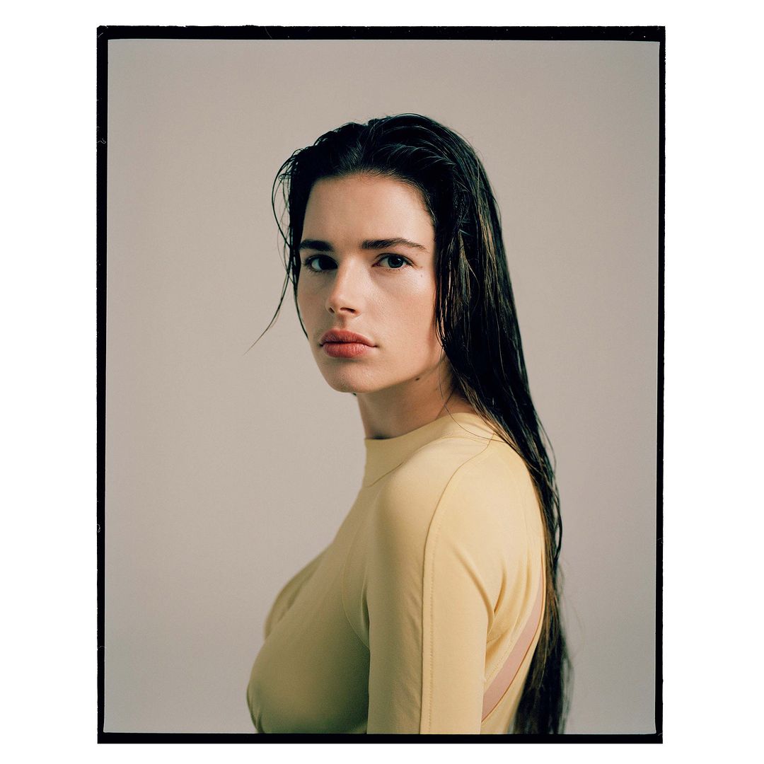 Portrait of a woman with long dark wet hair wearing a yellow top against a plain studio background.