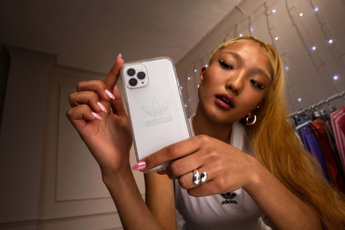 Young woman poses indoors holding a white smartphone with an Adidas logo on the case, wearing a white shirt and hoop earrings.