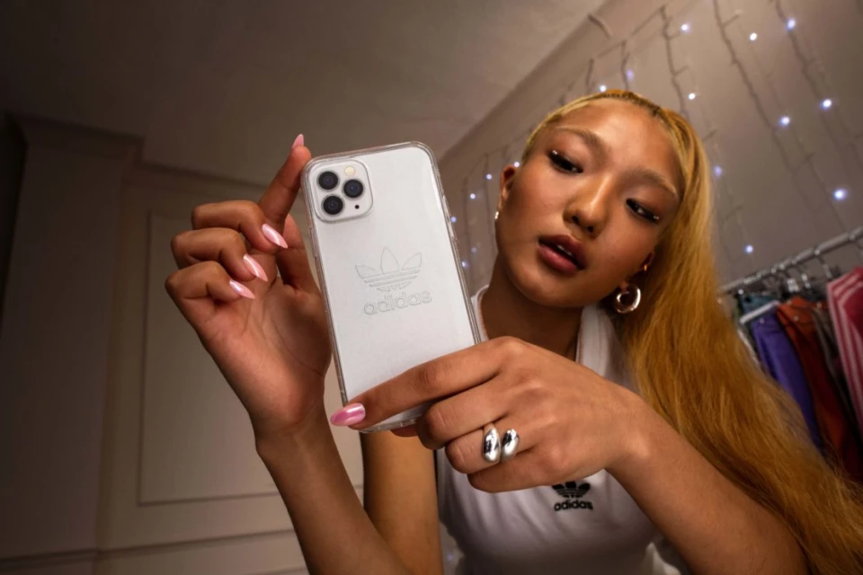 Young woman poses indoors holding a white smartphone with an Adidas logo on the case, wearing a white shirt and hoop earrings.