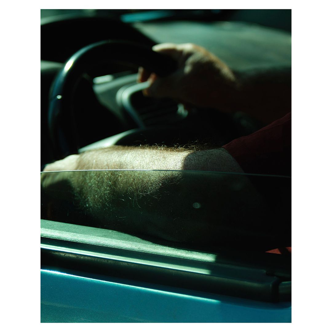 A man's arm rests on a car door with his hand on the steering wheel. The scene is cast in a warm, slightly moody light.