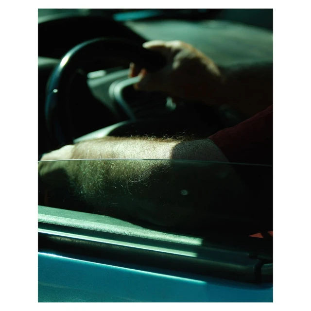 A man's arm rests on a car door with his hand on the steering wheel. The scene is cast in a warm, slightly moody light.