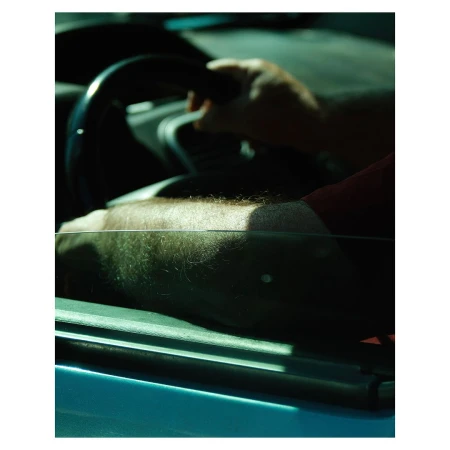 A man's arm rests on a car door with his hand on the steering wheel. The scene is cast in a warm, slightly moody light.