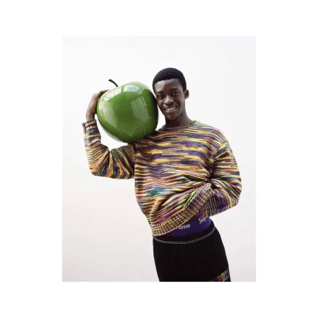 Black man holding a large glossy green apple prop wearing a multicolored striped sweater against white seamless