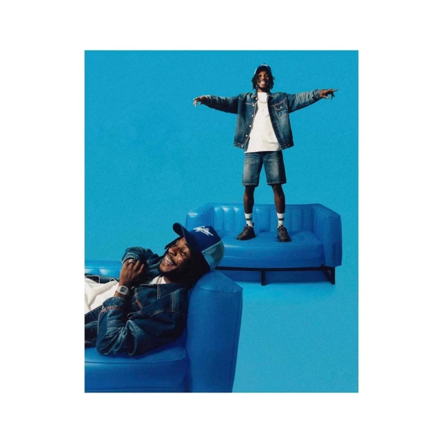 Two men pose in denim outfits on blue couches against a blue backdrop, one lying down, the other standing with arms outstretched.