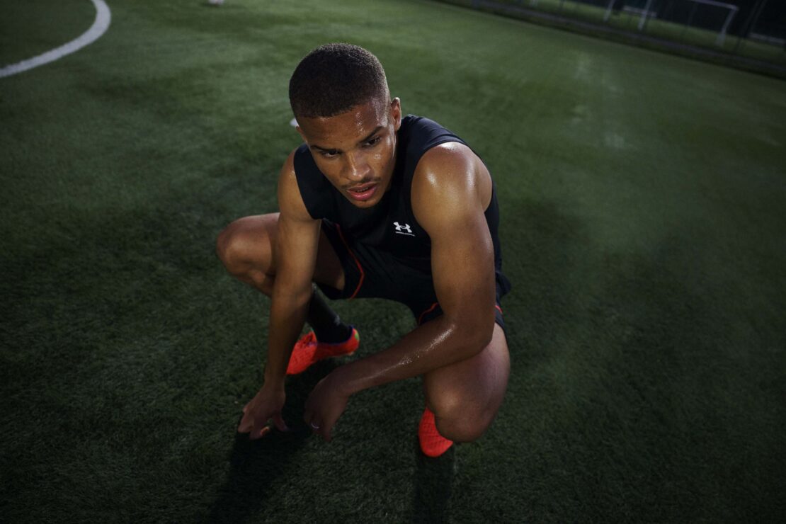 Young adult male athlete in black sleeveless athletic top, kneeling on green turf field in an outdoor sports setting at nighttime.