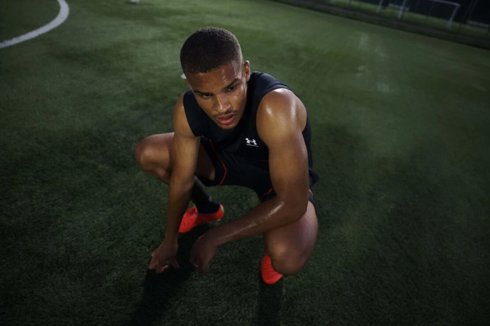Young adult male athlete in black sleeveless athletic top, kneeling on green turf field in an outdoor sports setting at nighttime.