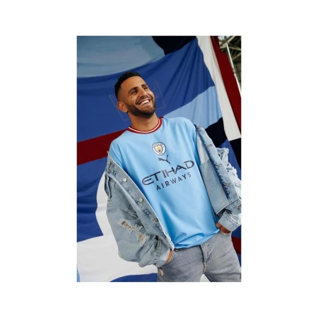 A man smiles wearing a Manchester City soccer jersey, denim jacket, and jeans, with a blue-and-white striped flag in the background.