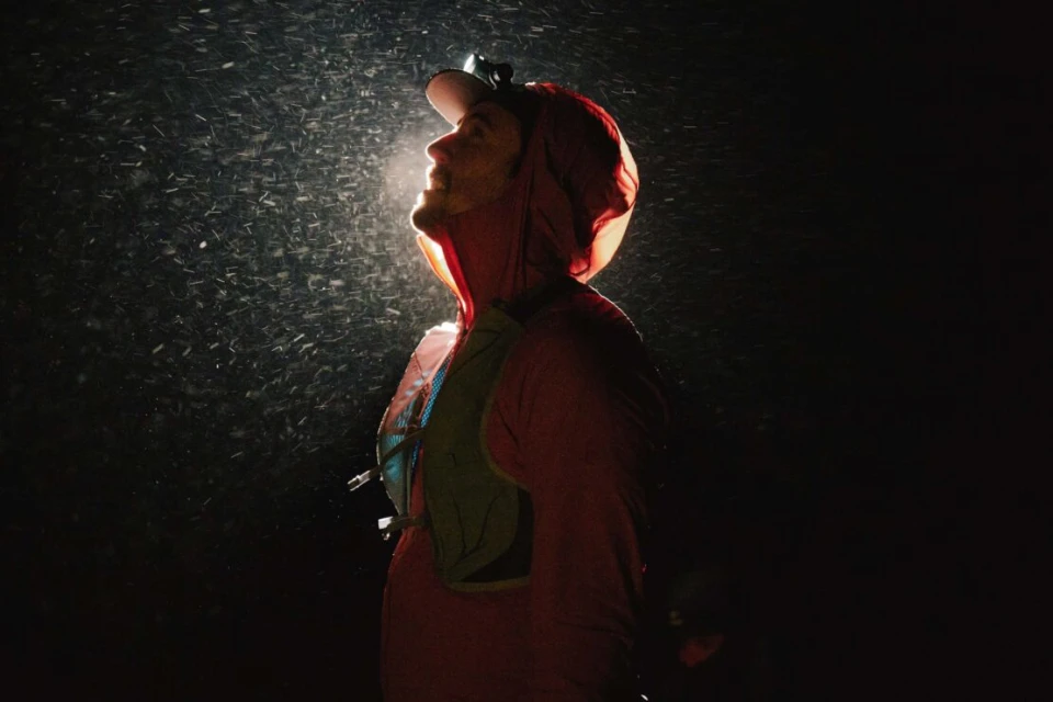 A person wearing a hooded jacket and headlamp looks up into falling snow against a dark background, illuminated by the headlamp.