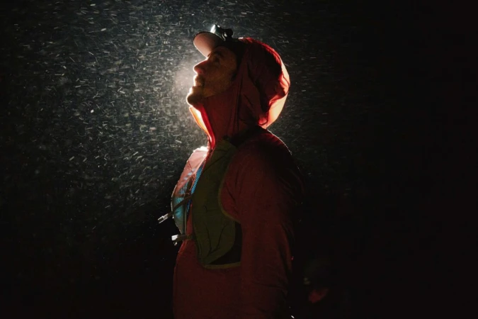 A person wearing a hooded jacket and headlamp looks up into falling snow against a dark background, illuminated by the headlamp.