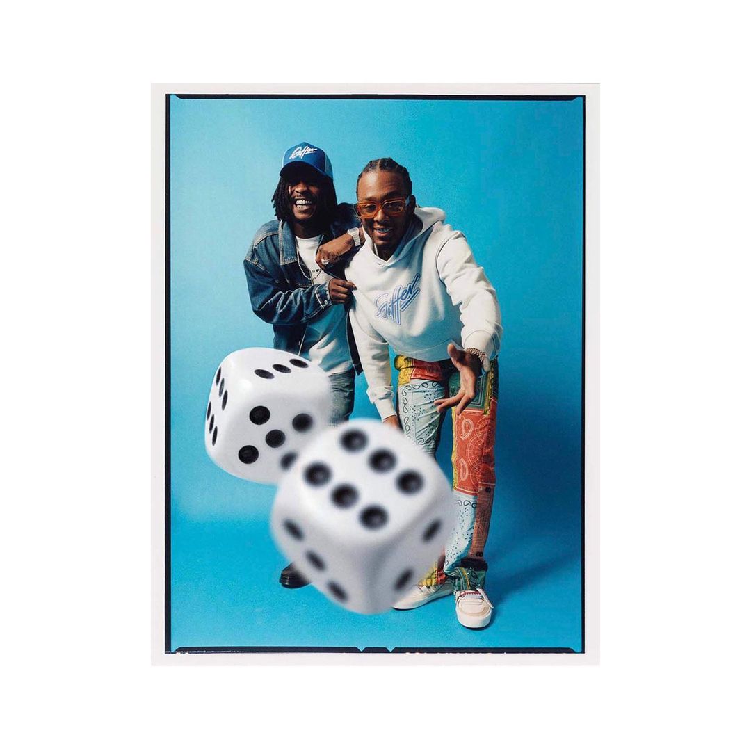 Two smiling men pose in front of a light blue backdrop with floating dice, wearing streetwear, with a medium shot and indoor lighting.
