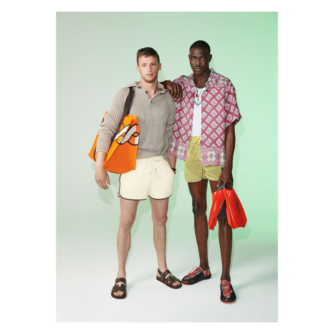 Two men pose in a brightly lit studio wearing summer outfits, one carrying a tote bag and the other holding red swim fins.