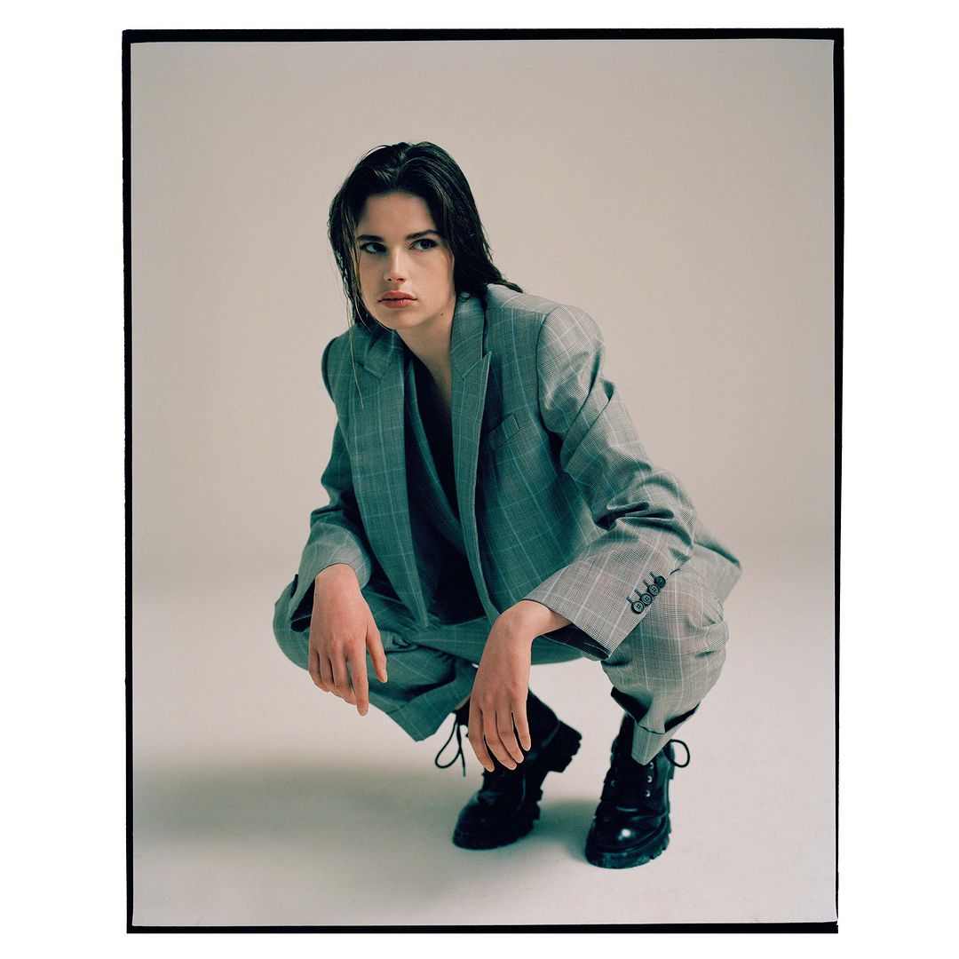 Woman in a grey plaid suit crouching in a studio, looking away with wet hair and black boots in a fashion style editorial shot.