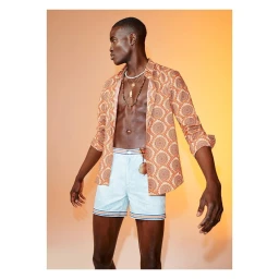 Black man wearing an unbuttoned patterned orange shirt and light blue shorts against a warm gradient background.