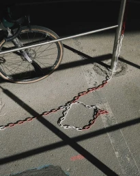 Bicycle wheel secured with a red and white chain to a metal post casting sharp diagonal shadows on asphalt pavement.