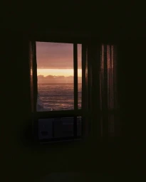 View of the ocean at sunset through a dark interior window framed by curtains