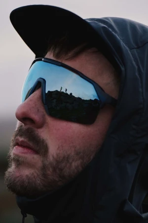 Close-up portrait of a man wearing reflective sunglasses and a dark hooded jacket outdoors.
