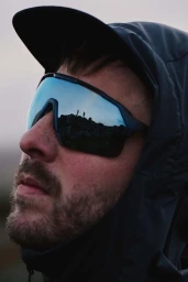 Close-up portrait of a man wearing reflective sunglasses and a dark hooded jacket outdoors.
