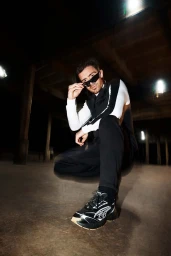 Man wearing dark sunglasses and layered sportswear crouching beneath a concrete structure at night