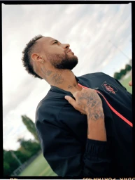 Portrait of Neymar Jr wearing a black jacket outdoors against a pale sky and blurred green background, photographed by Elliott Wilcox.